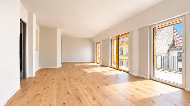 Helles, leeres Wohnzimmer mit Holzboden, großen Fenstern und Zugang zu einem Balkon.