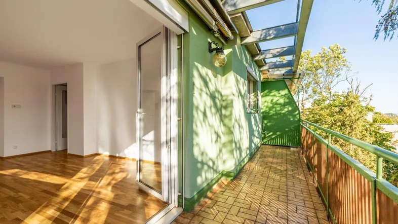 Helles Wohnzimmer mit Parkettboden und Zugang zu einem langen Balkon mit grüner Fassade.