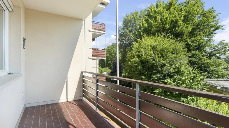 Geräumiger Balkon mit Fliesenboden und Holzbalkongeländer, umgeben von viel Grün und Bäumen.