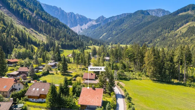 Luftaufnahme eines Hauses in einem malerischen Tal, umgeben von Wäldern und hohen Bergen.