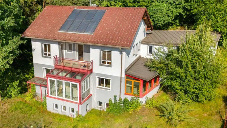 Freistehendes Einfamilienhaus mit rotem Ziegeldach, Solaranlage und Wintergarten, umgeben von viel Grün.