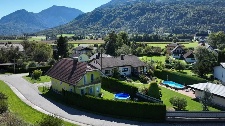 Luftaufnahme eines Wohngebiets mit Häusern, Gärten und einem Pool vor einer malerischen Bergkulisse.
