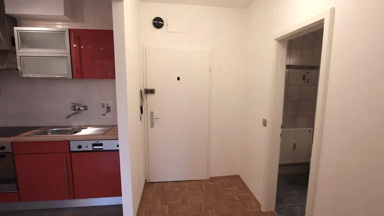 Heller Eingangsbereich mit Parkettboden, der zu einer roten Küche und einem Badezimmer führt.