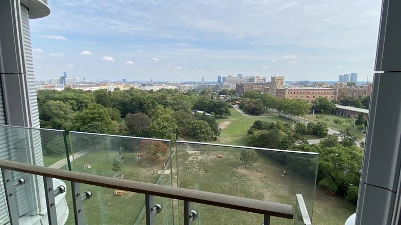 Panoramablick vom Balkon über einen grünen Park und die Stadtlandschaft unter einem bewölkten Himmel.