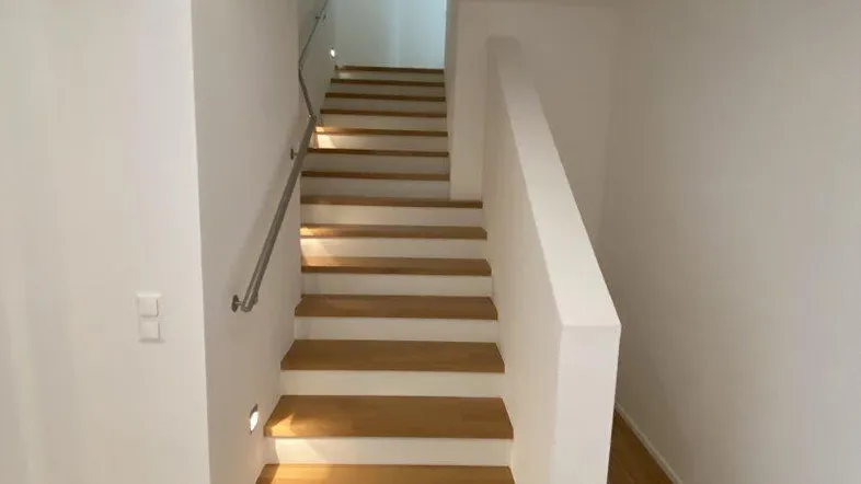 Moderne Holztreppe mit Edelstahlgeländer und integrierter Beleuchtung in einem hellen Flur.