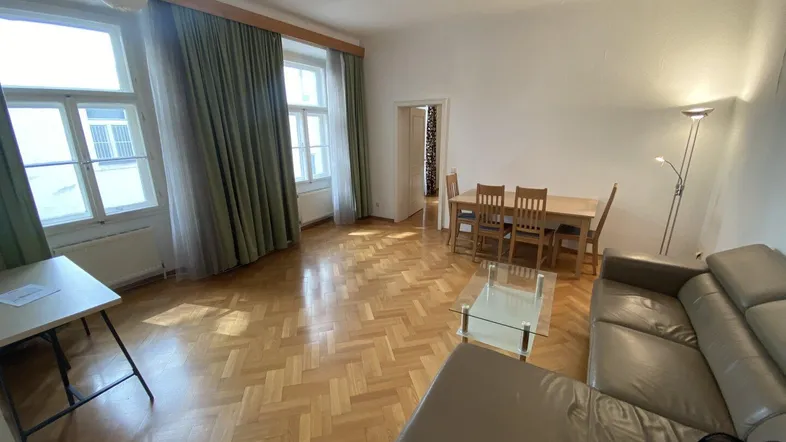 Heller Wohn-Essbereich mit Fischgrätparkett, beigem Ledersofa und großzügigen Fenstern mit grünen Vorhängen.