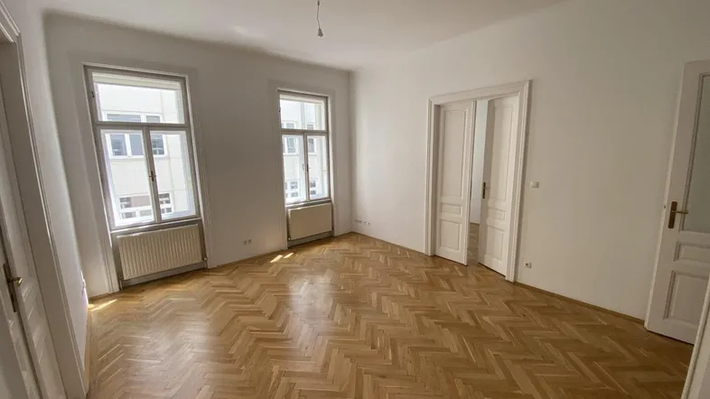Geräumiges Wohnzimmer mit Fischgrätparkett und großen Fenstern, die viel Tageslicht hereinlassen.
