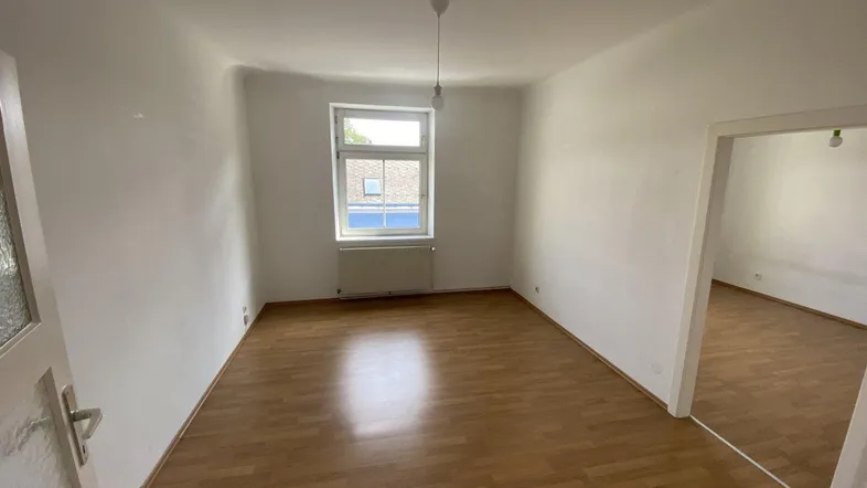 Helles, leeres Zimmer mit Laminatboden und einem Fenster, das Tageslicht hereinlässt.