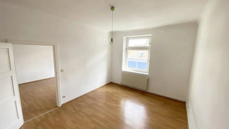 Heller, leerer Raum mit Laminatboden, weißen Wänden und einem Fenster mit Heizkörper.