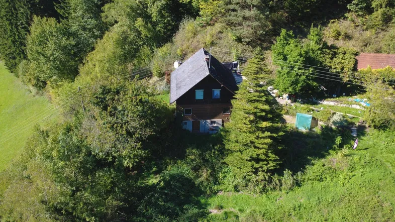 Luftaufnahme eines abgelegenen Hauses mit dunklem Dach, umgeben von dichtem Wald und grünen Wiesen.