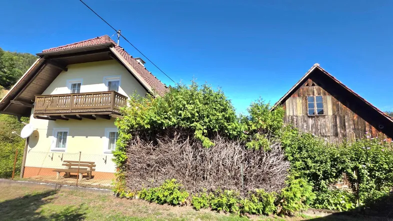 Ein traditionelles Haus mit Holzbalkon und einem Nebengebäude, umgeben von üppigem Grün unter blauem Himmel.