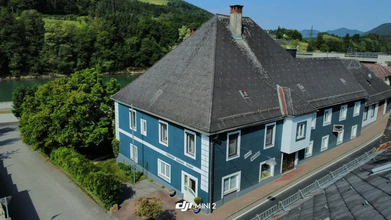 Luftaufnahme eines historischen Gebäudes mit blauer Fassade, umgeben von grünen Hügeln und einem See.