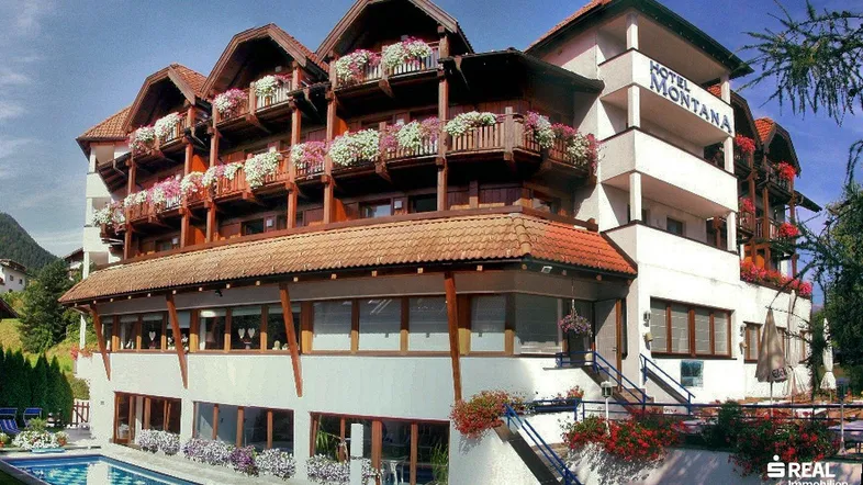 Hotelgebäude im alpinen Stil mit blühenden Balkonen und Außenpool unter blauem Himmel.