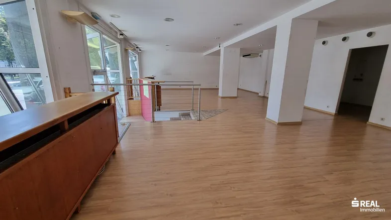 Großer, heller Innenraum mit Laminatboden, hohen Fenstern und einer Treppe zum Untergeschoss.