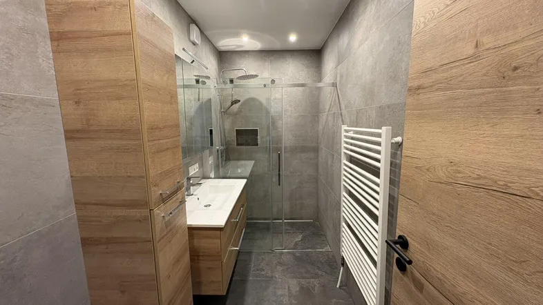 Modernes Badezimmer mit begehbarer Dusche, Holzschränken, Einzelwaschbecken und grauen Fliesen.