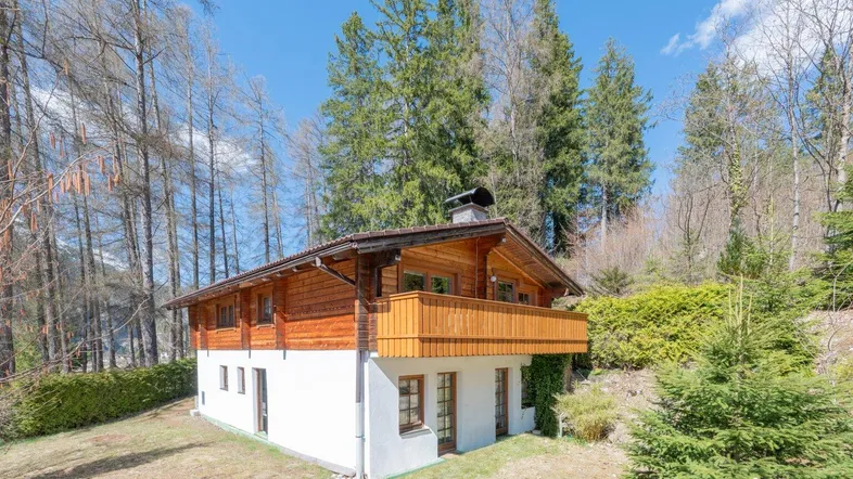 Traditionelles Holzhaus mit weißem Sockel und Balkon, umgeben von Bäumen in alpiner Landschaft.