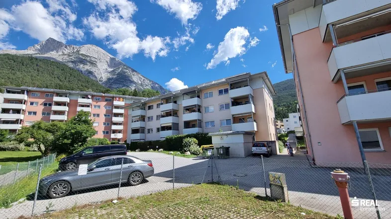 Mehrere Wohngebäude mit Bergkulisse und Parkplätzen unter blauem Himmel.