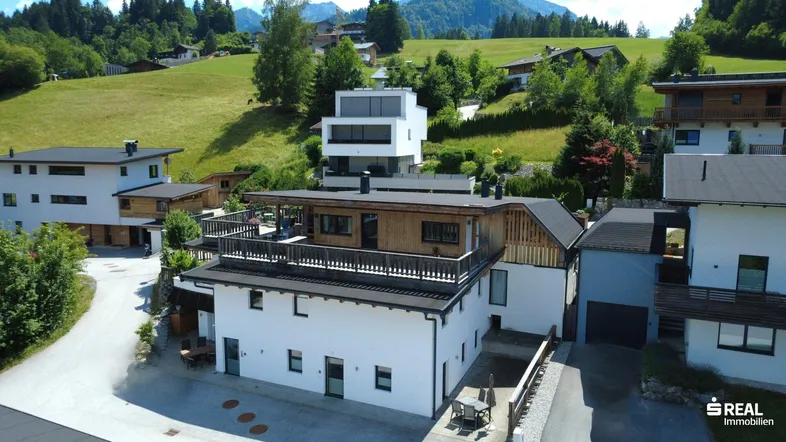 Luftaufnahme eines modernen Wohnhauses mit mehreren Terrassen in alpiner Landschaft.