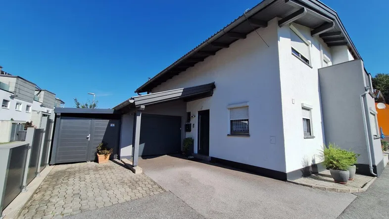 Modernes Einfamilienhaus mit weißer Fassade, Carport und gepflasterter Auffahrt unter blauem Himmel.