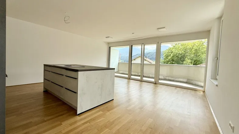 Heller offener Wohn-Essbereich mit moderner Kücheninsel, Holzboden und Zugang zu einem Balkon mit Bergblick.