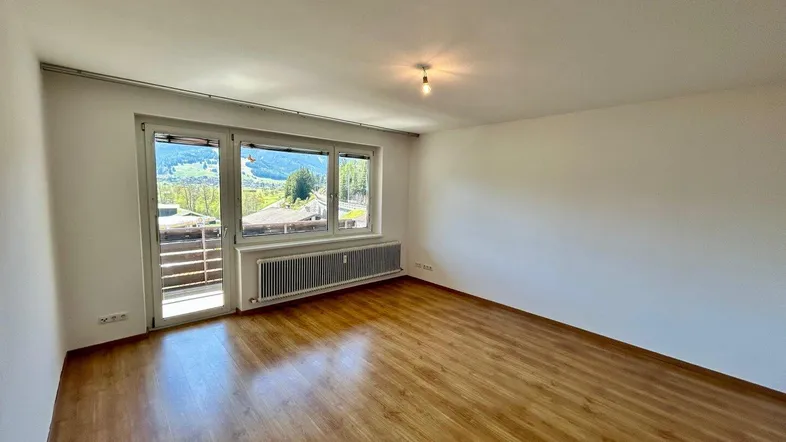 Helles Wohnzimmer mit Laminatboden, großem Fenster und Zugang zum Balkon mit Bergblick.