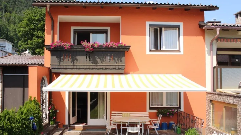 Hausfassade mit Terrasse, Markise und Balkon, umgeben von grüner Natur und Bergen.