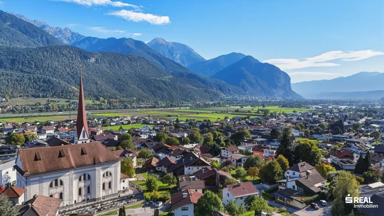 Weitläufige Luftaufnahme einer Stadt mit Kirche, umgeben von grünen Feldern und majestätischen Bergen.