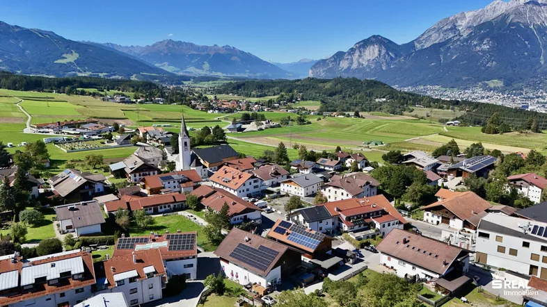 Luftaufnahme eines malerischen Dorfes mit Kirche, umgeben von grünen Feldern und majestätischen Bergen.
