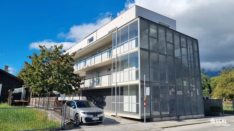 Modernes Mehrfamilienhaus mit Glasfassade und Balkonen, davor ein geparktes Auto auf dem Stellplatz.