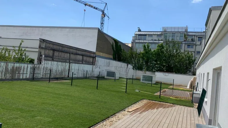 Großer Gartenbereich mit Rasenfläche und Holzterrasse, umgeben von einem Zaun und Gebäuden im Hintergrund.