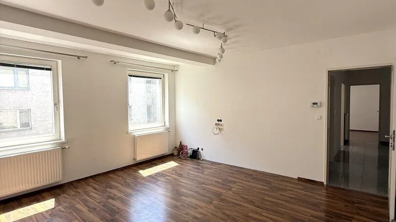 Helles Wohnzimmer mit dunklem Laminatboden und zwei Fenstern, bereit zur individuellen Einrichtung.