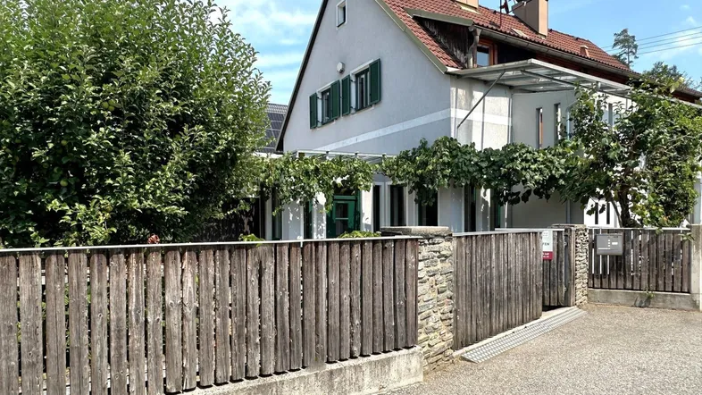 Traditionelles Haus mit Holzzaun, grünen Fensterläden und bewachsenem Eingangsbereich.