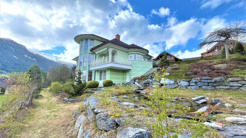Großzügiges Haus mit runder Architektur, Balkon und angelegtem Steingarten vor alpiner Kulisse.