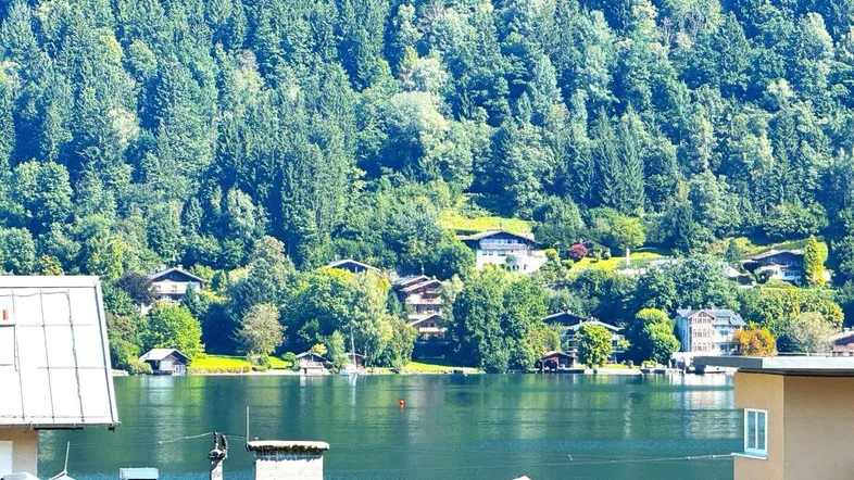 Panoramablick auf den See und die umliegenden bewaldeten Berge, teilweise sichtbar sind Dächer mit Solaranlagen.