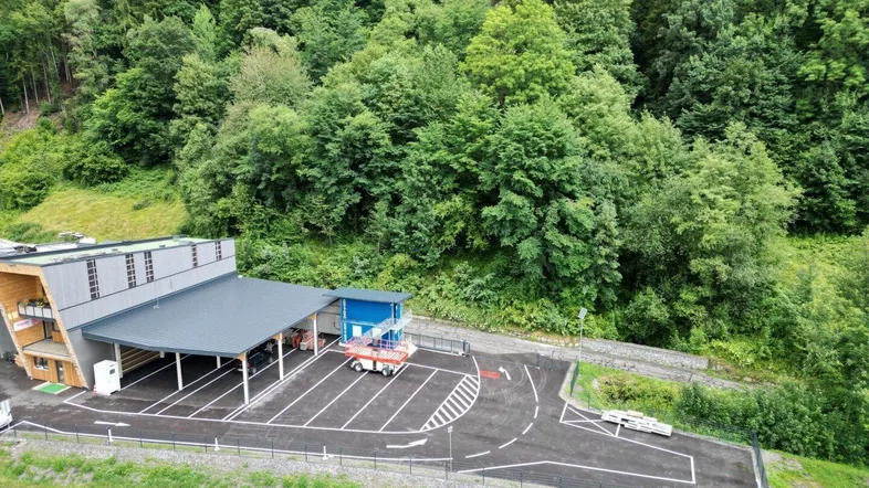 Luftaufnahme eines Gewerbegebäudes mit großem Parkplatz, umgeben von dichtem Wald und Bergen.