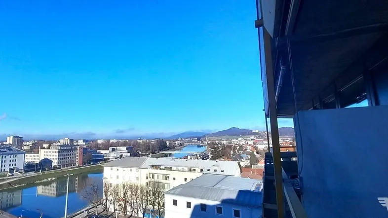 Weitläufiger Stadtblick von oben mit Fluss, Gebäuden und Bergen am Horizont unter blauem Himmel.