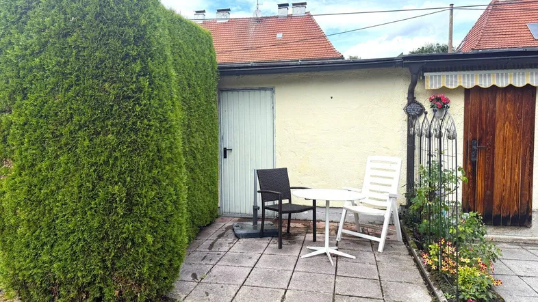 Gemütliche gepflasterte Terrasse mit Sitzgelegenheiten, umgeben von einer hohen Hecke und Blumenbeeten.