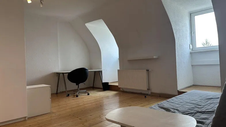 Dachgeschosszimmer mit schrägen Wänden, Holzboden und einem Fenster, das viel natürliches Licht hereinlässt.
