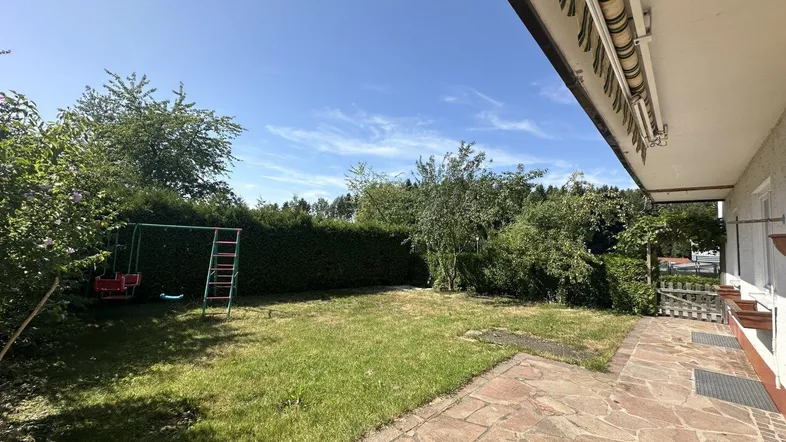 Sonniger Gartenbereich mit Rasenfläche, Hecke, Spielgeräten und gepflasterter Terrasse.