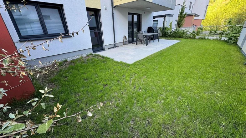Gepflegter Gartenbereich mit Rasenfläche und einer möblierten Terrasse vor einem modernen Gebäude.