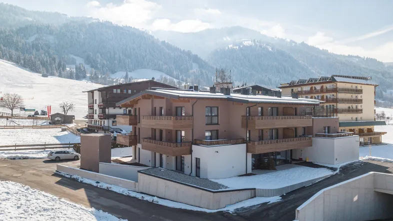 Modernes Apartmentgebäude in alpiner Winterlandschaft mit verschneiten Bergen im Hintergrund.