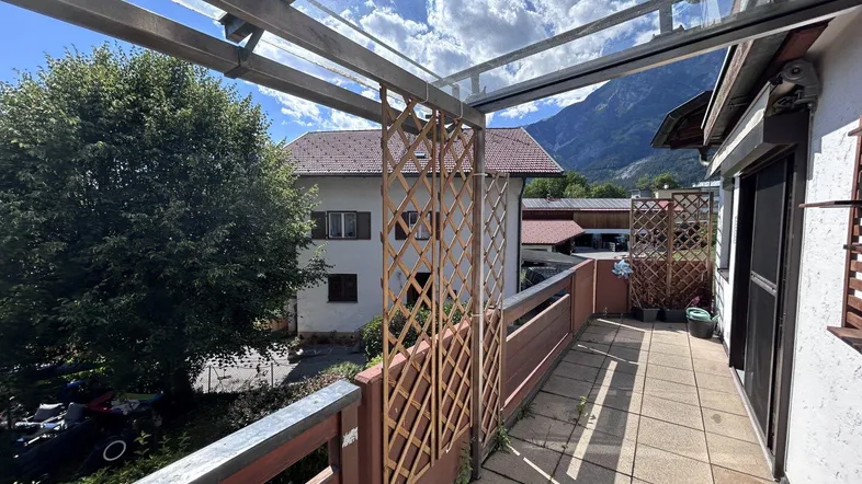 Geräumiger Balkon mit Glasüberdachung, Holzelementen und Bergblick unter blauem Himmel.