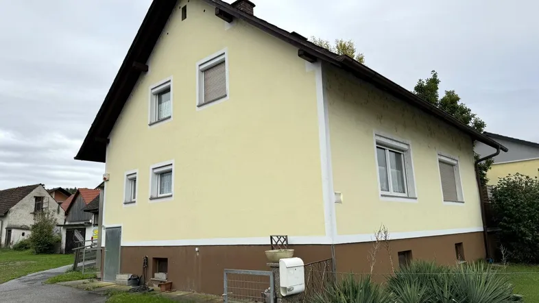 Gelb gestrichenes Einfamilienhaus mit braunem Sockel und Rollläden unter bewölktem Himmel.