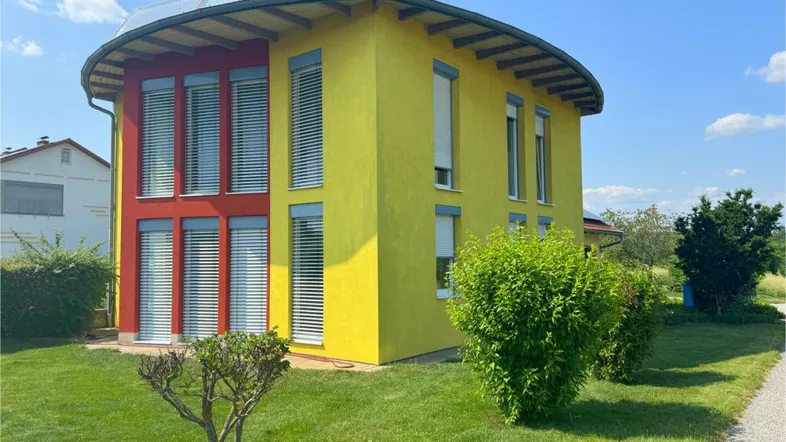 Modernes Haus mit gelber und roter Fassade, geschwungenem Dach und Photovoltaikanlage auf grünem Rasen.