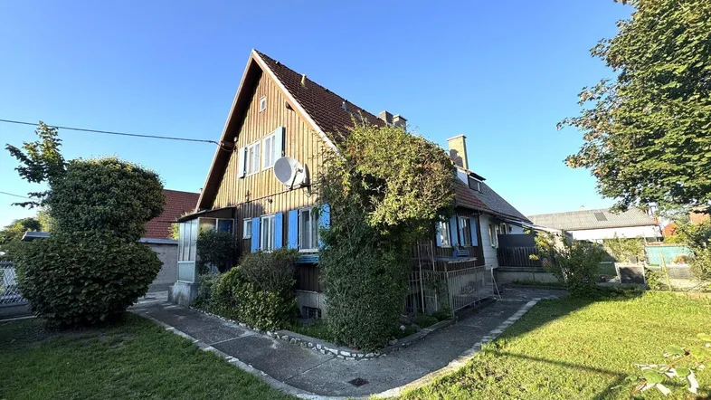 Hausfassade mit Holzverkleidung, Satteldach und gepflegtem Vorgarten unter blauem Himmel.