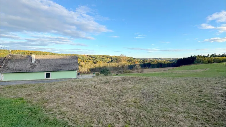 Blick auf ein ländliches Haus mit grüner Fassade, umgeben von Feldern und Wäldern unter blauem Himmel.