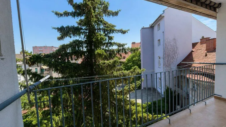Geräumiger Balkon mit Fliesenboden und Metallgeländer, umgeben von hohen Bäumen und Gebäuden.