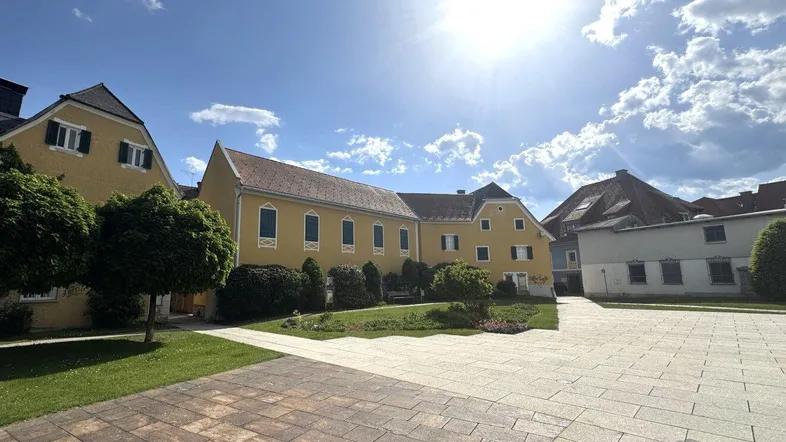 Gelbes Gebäude mit gepflegtem Rasen und gepflastertem Vorplatz unter blauem Himmel.