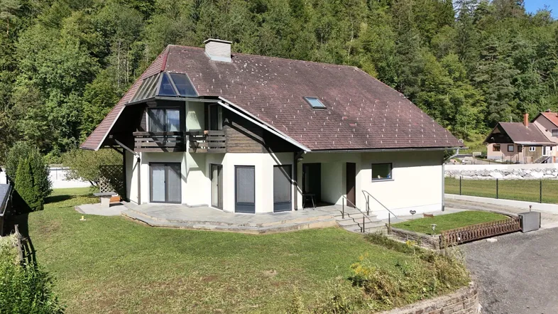 Hausfassade mit Satteldach, Balkon und Terrasse, umgeben von grünen Hügeln.