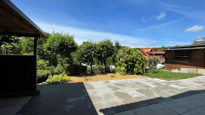 Große gepflasterte Terrasse mit Blick auf den grünen Garten und die umliegende Natur unter blauem Himmel.